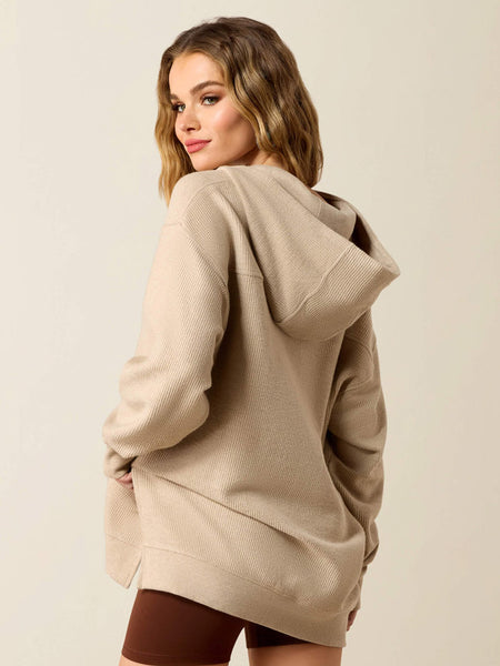 A woman wearing an oversized beige waffle-knit hoodie, photographed from the back and side against a plain light beige studio background. If you need any assistance with this item or the purchase of this item please call us at five six one seven four eight eight eight zero one Monday through Saturday 10:00a.m EST to 8:00 p.m EST