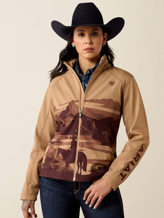 A woman wearing a tan zip-front jacket with a brown western mountain and rider graphic, a black cowboy hat, and dark blue jeans, photographed against a plain light beige studio background. The jacket has an ARIAT logo on the chest and ARIAT text down the sleeve. If you need any assistance with this item or the purchase of this item please call us at five six one seven four eight eight eight zero one Monday through Saturday 10:00a.m EST to 8:00 p.m EST