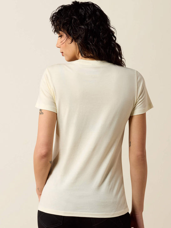A woman with curly dark hair is shown from the back wearing a short-sleeve cream-colored t-shirt and black pants, set against a plain light beige studio background. If you need any assistance with this item or the purchase of this item please call us at five six one seven four eight eight eight zero one Monday through Saturday 10:00a.m EST to 8:00 p.m EST