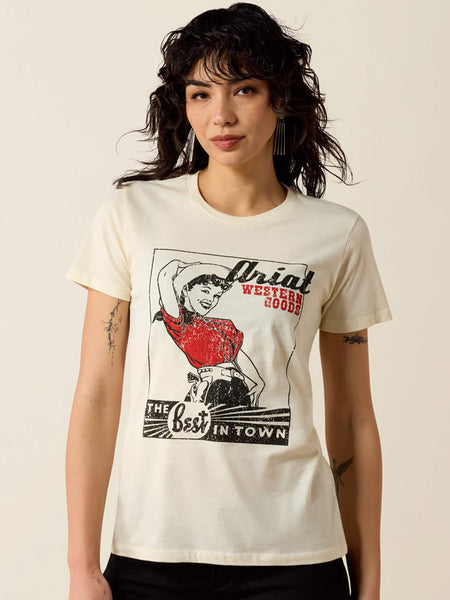 A woman wearing a cream-colored graphic t-shirt with a vintage-style western print featuring a woman in a red dress and the text 'Ariat Western Goods'. If you need any assistance with this item or the purchase of this item please call us at five six one seven four eight eight eight zero one Monday through Saturday 10:00a.m EST to 8:00 p.m EST