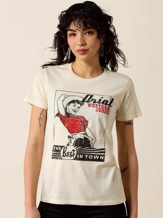 A woman wearing a cream-colored graphic t-shirt with a vintage-style western print featuring a woman in a red dress and the text 'Ariat Western Goods'. If you need any assistance with this item or the purchase of this item please call us at five six one seven four eight eight eight zero one Monday through Saturday 10:00a.m EST to 8:00 p.m EST