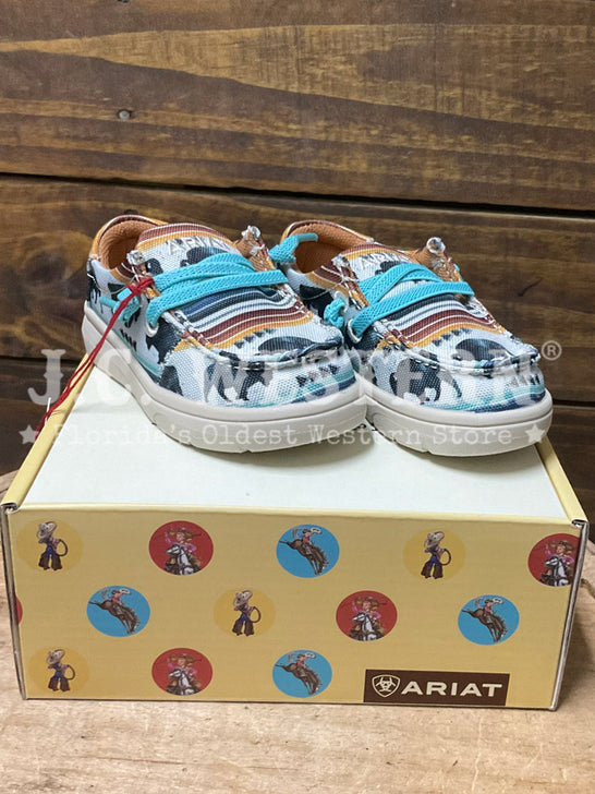 A pair of Ariat slip-on western-style shoes with a multicolor cowboy print and turquoise laces, displayed on top of their yellow branded box against a rustic wooden background. If you need any assistance with this item or the purchase of this item please call us at five six one seven four eight eight eight zero one Monday through Saturday 10:00a.m EST to 8:00 p.m EST