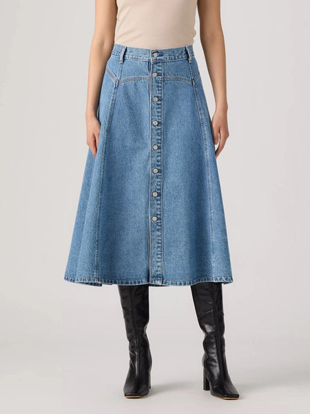 A model wearing a high-waisted, light blue denim A-line midi skirt with a button-down front, paired with a beige top and black leather knee-high boots, against a light gray studio background. If you need any assistance with this item or the purchase of this item please call us at five six one seven four eight eight eight zero one Monday through Saturday 10:00a.m EST to 8:00 p.m EST