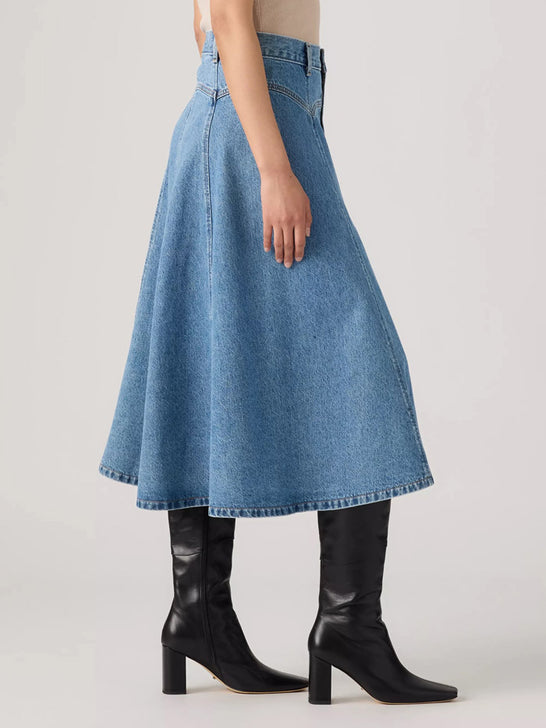 A model wearing a high-waisted, flared blue denim midi skirt paired with black leather knee-high heeled boots, shown from the waist down against a light gray studio background. If you need any assistance with this item or the purchase of this item please call us at five six one seven four eight eight eight zero one Monday through Saturday 10:00a.m EST to 8:00 p.m EST