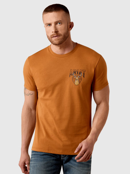 A man wearing a burnt orange crewneck t-shirt with a distressed graphic on the left chest featuring the word 'ARIAT' and a horned mask, paired with blue jeans, against a light gray background. If you need any assistance with this item or the purchase of this item please call us at five six one seven four eight eight eight zero one Monday through Saturday 10:00a.m EST to 8:00 p.m EST