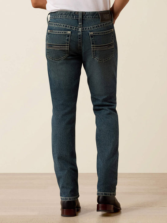 Rear view of a person wearing dark blue slim-fit jeans with contrast stitching and a brown leather patch on the back waistband, standing against a plain beige studio background. If you need any assistance with this item or the purchase of this item please call us at five six one seven four eight eight eight zero one Monday through Saturday 10:00a.m EST to 8:00 p.m EST