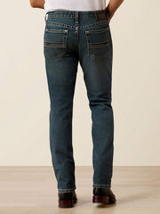 Rear view of a person wearing dark blue slim-fit jeans with contrast stitching and a brown leather patch on the back waistband, standing against a plain beige studio background. If you need any assistance with this item or the purchase of this item please call us at five six one seven four eight eight eight zero one Monday through Saturday 10:00a.m EST to 8:00 p.m EST