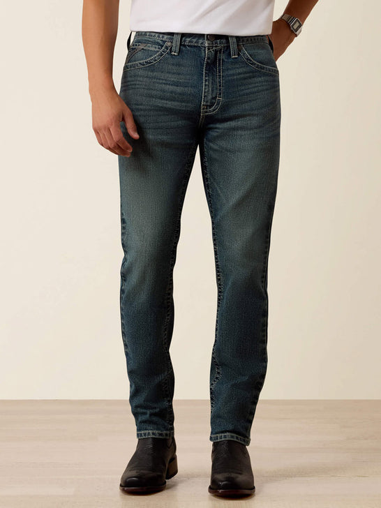 A person wearing dark blue slim-fit jeans with a white t-shirt and black boots, standing against a light beige studio background. If you need any assistance with this item or the purchase of this item please call us at five six one seven four eight eight eight zero one Monday through Saturday 10:00a.m EST to 8:00 p.m EST