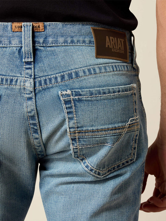 Close-up back view of a person wearing light blue denim jeans, showing the waistband, belt loops, back pocket, and Ariat brand patch. If you need any assistance with this item or the purchase of this item please call us at five six one seven four eight eight eight zero one Monday through Saturday 10:00a.m EST to 8:00 p.m EST