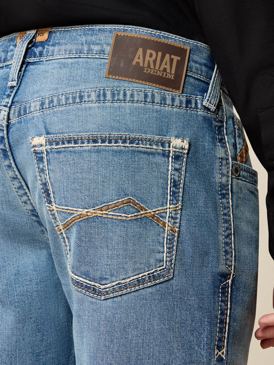 Close-up of the back waistband and back pocket of light blue denim jeans, showing a brown Ariat Denim brand patch and contrast stitching. If you need any assistance with this item or the purchase of this item please call us at five six one seven four eight eight eight zero one Monday through Saturday 10:00a.m EST to 8:00 p.m EST