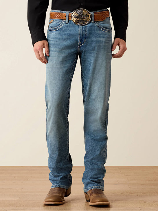 A person wearing light blue denim jeans with a brown woven belt and a large decorative silver buckle, paired with a black long-sleeve shirt and brown boots, standing against a plain beige wall and light wood floor. If you need any assistance with this item or the purchase of this item please call us at five six one seven four eight eight eight zero one Monday through Saturday 10:00a.m EST to 8:00 p.m EST