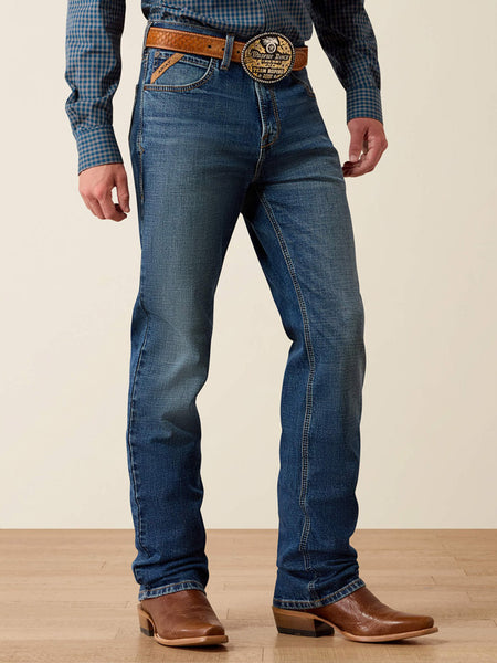 A person wearing blue denim jeans with a brown leather belt and a large decorative buckle, paired with brown cowboy boots and a blue plaid shirt, standing against a plain beige wall and light wood floor. If you need any assistance with this item or the purchase of this item please call us at five six one seven four eight eight eight zero one Monday through Saturday 10:00a.m EST to 8:00 p.m EST