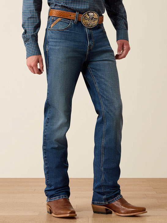 A person wearing blue denim jeans with a brown leather belt and a large decorative buckle, paired with brown cowboy boots and a blue plaid shirt, standing against a plain beige wall and light wood floor. If you need any assistance with this item or the purchase of this item please call us at five six one seven four eight eight eight zero one Monday through Saturday 10:00a.m EST to 8:00 p.m EST