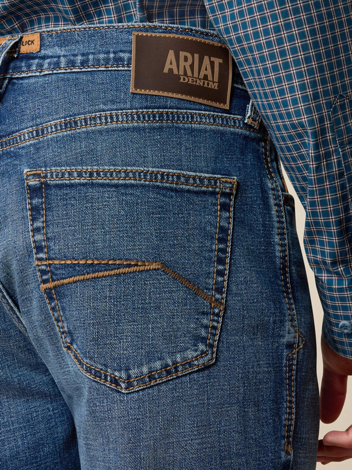 A person wearing blue denim jeans with a brown leather belt and a large decorative buckle, paired with brown cowboy boots and a blue plaid shirt, standing against a plain beige wall and light wood floor. If you need any assistance with this item or the purchase of this item please call us at five six one seven four eight eight eight zero one Monday through Saturday 10:00a.m EST to 8:00 p.m EST