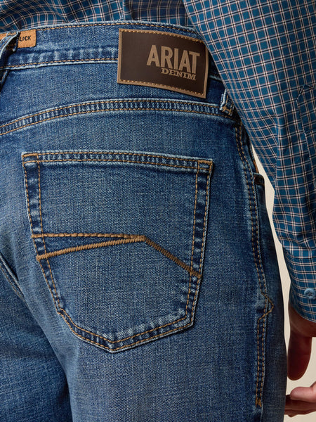 Close-up of the back waistband and pocket area of blue denim jeans, showing the Ariat Denim brand patch and a blue plaid shirt tucked into the waistband. If you need any assistance with this item or the purchase of this item please call us at five six one seven four eight eight eight zero one Monday through Saturday 10:00a.m EST to 8:00 p.m EST