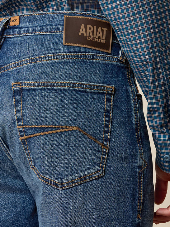 Close-up of the back waistband and pocket area of blue denim jeans, showing the Ariat Denim brand patch and a blue plaid shirt tucked into the waistband. If you need any assistance with this item or the purchase of this item please call us at five six one seven four eight eight eight zero one Monday through Saturday 10:00a.m EST to 8:00 p.m EST