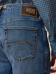 Close-up of the back waistband and pocket area of blue denim jeans, showing the Ariat Denim brand patch and a blue plaid shirt tucked into the waistband. If you need any assistance with this item or the purchase of this item please call us at five six one seven four eight eight eight zero one Monday through Saturday 10:00a.m EST to 8:00 p.m EST