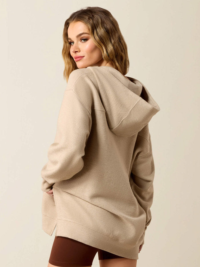 A woman wearing a beige knit pullover hoodie with a split neckline and a small embroidered logo on the left chest, paired with dark brown shorts, against a plain light beige background. If you need any assistance with this item or the purchase of this item please call us at five six one seven four eight eight eight zero one Monday through Saturday 10:00a.m EST to 8:00 p.m EST