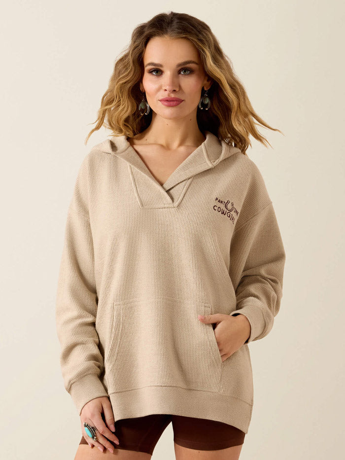 A woman wearing a beige knit pullover hoodie with a split neckline and a small embroidered logo on the left chest, paired with dark brown shorts, against a plain light beige background. If you need any assistance with this item or the purchase of this item please call us at five six one seven four eight eight eight zero one Monday through Saturday 10:00a.m EST to 8:00 p.m EST