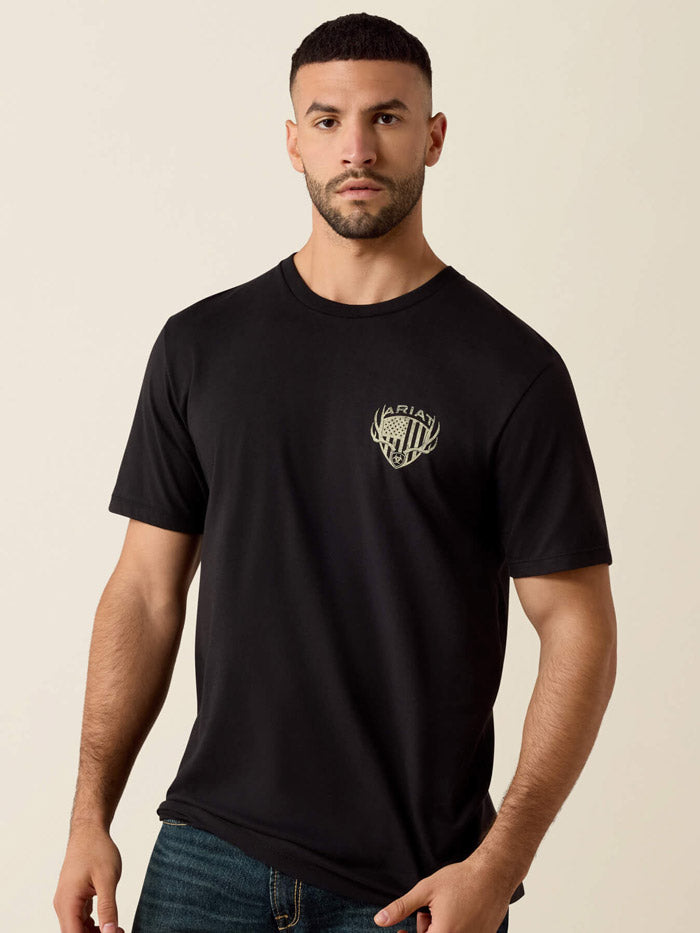 A man viewed from the back wearing a black short-sleeve T-shirt with a large distressed off-white graphic on the back featuring the word ARIAT, antlers, a shield, and an American flag motif. If you need any assistance with this item or the purchase of this item please call us at five six one seven four eight eight eight zero one Monday through Saturday 10:00a.m EST to 8:00 p.m EST