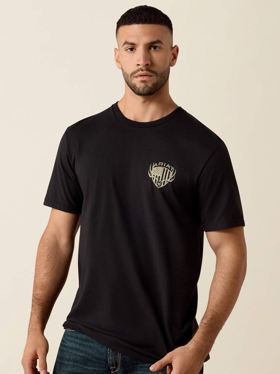 A man wearing a black short-sleeve crewneck t-shirt with a small beige graphic on the left chest, paired with blue jeans, against a solid light beige background. If you need any assistance with this item or the purchase of this item please call us at five six one seven four eight eight eight zero one Monday through Saturday 10:00a.m EST to 8:00 p.m EST