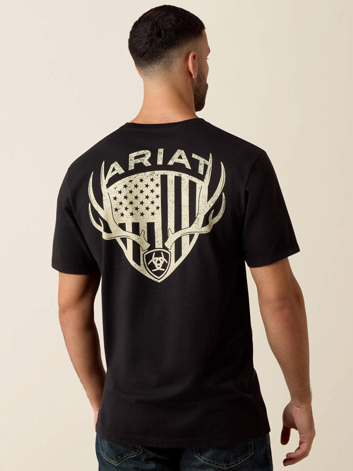 A man viewed from the back wearing a black short-sleeve T-shirt with a large distressed off-white graphic on the back featuring the word ARIAT, antlers, a shield, and an American flag motif. If you need any assistance with this item or the purchase of this item please call us at five six one seven four eight eight eight zero one Monday through Saturday 10:00a.m EST to 8:00 p.m EST