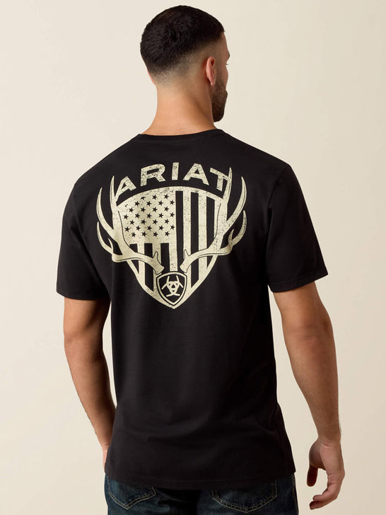 A man viewed from the back wearing a black short-sleeve T-shirt with a large distressed off-white graphic on the back featuring the word ARIAT, antlers, a shield, and an American flag motif. If you need any assistance with this item or the purchase of this item please call us at five six one seven four eight eight eight zero one Monday through Saturday 10:00a.m EST to 8:00 p.m EST