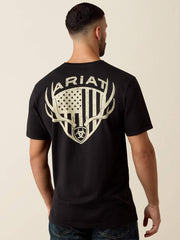 A man viewed from the back wearing a black short-sleeve T-shirt with a large distressed off-white graphic on the back featuring the word ARIAT, antlers, a shield, and an American flag motif. If you need any assistance with this item or the purchase of this item please call us at five six one seven four eight eight eight zero one Monday through Saturday 10:00a.m EST to 8:00 p.m EST