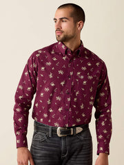 A man wearing a long-sleeve button-down shirt in burgundy with a beige and white cowboy motif, paired with dark gray jeans and a silver belt, against a solid light beige background. If you need any assistance with this item or the purchase of this item please call us at five six one seven four eight eight eight zero one Monday through Saturday 10:00a.m EST to 8:00 p.m EST