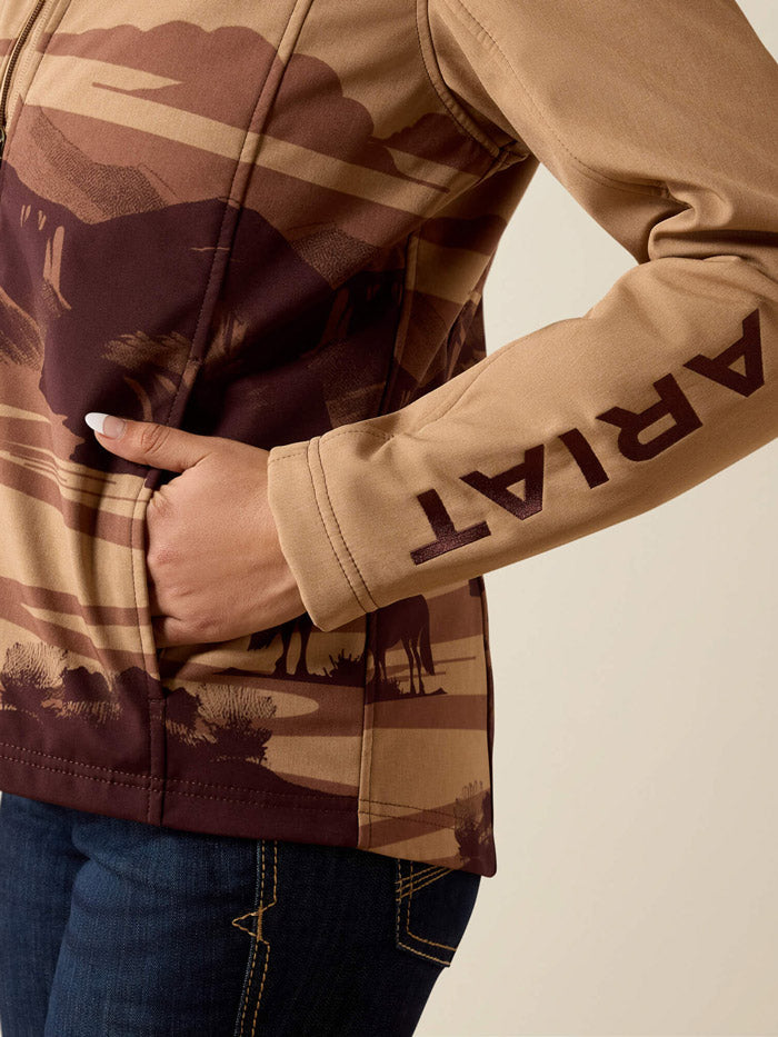 A woman wearing a tan zip-front jacket with a brown western mountain and rider graphic, a black cowboy hat, and dark blue jeans, photographed against a plain light beige studio background. The jacket has an ARIAT logo on the chest and ARIAT text down the sleeve. If you need any assistance with this item or the purchase of this item please call us at five six one seven four eight eight eight zero one Monday through Saturday 10:00a.m EST to 8:00 p.m EST