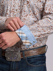 A close-up of a man's hands adjusting the cuff of a long-sleeve western shirt. The shirt features a brown, beige, and blue paisley pattern, a light blue textured cuff, and silver snap buttons. If you need any assistance with this item or the purchase of this item please call us at five six one seven four eight eight eight zero one Monday through Saturday 10:00a.m EST to 8:00 p.m EST