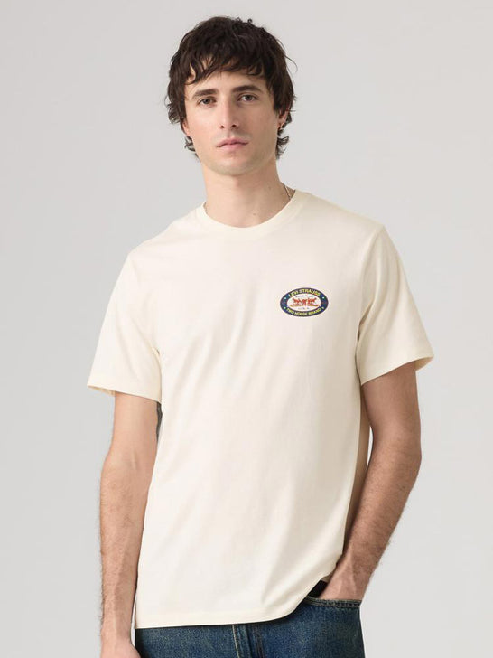 A man wearing a cream-colored short-sleeve t-shirt with a small circular 'WILD WEST' graphic on the left chest, paired with blue jeans, against a solid light gray background. If you need any assistance with this item or the purchase of this item please call us at five six one seven four eight eight eight zero one Monday through Saturday 10:00a.m EST to 8:00 p.m EST