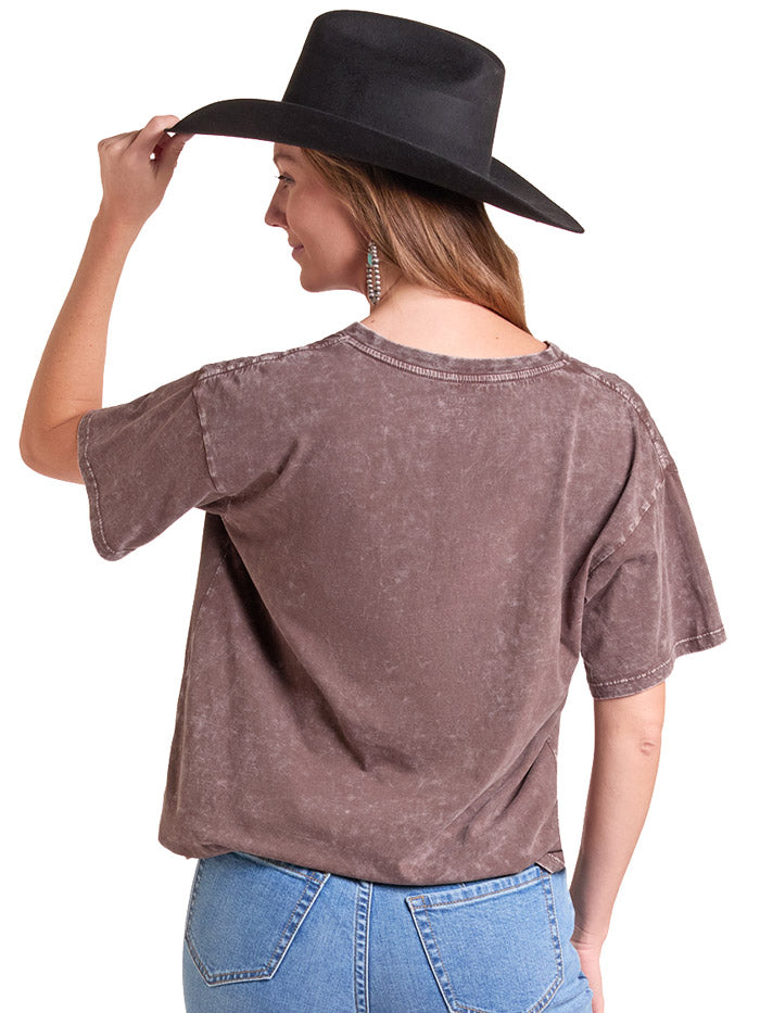 A woman wearing a brown acid-wash graphic t-shirt with a cow skull and 'Rodeo Cowboy' print, paired with a black cowboy hat and blue jeans, against a white background. If you need any assistance with this item or the purchase of this item please call us at five six one seven four eight eight eight zero one Monday through Saturday 10:00a.m EST to 8:00 p.m EST
