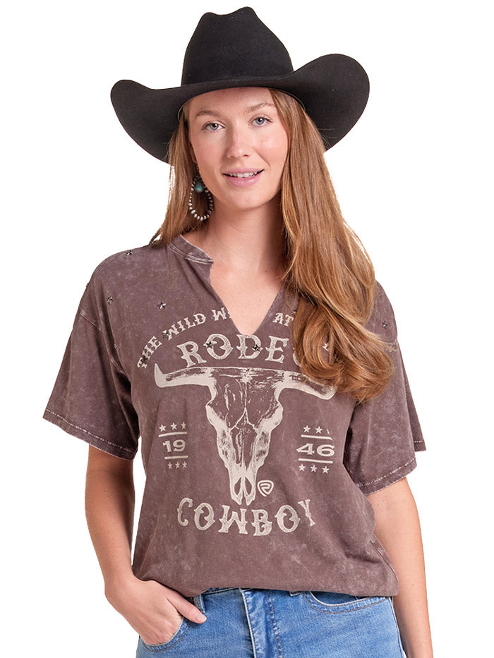 A woman wearing a brown acid-wash graphic t-shirt with a cow skull and 'Rodeo Cowboy' print, paired with a black cowboy hat and blue jeans, against a white background. If you need any assistance with this item or the purchase of this item please call us at five six one seven four eight eight eight zero one Monday through Saturday 10:00a.m EST to 8:00 p.m EST