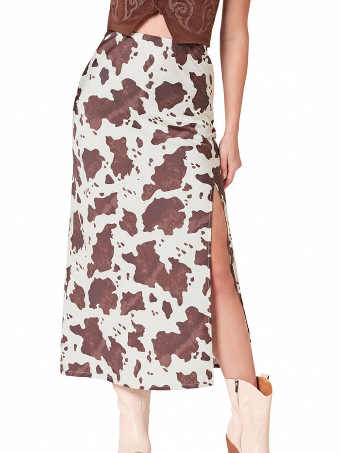 A model wearing a mid-length cow-print skirt with a high side slit, paired with a brown cropped top and beige cowboy boots, shown against a plain white background. If you need any assistance with this item or the purchase of this item please call us at five six one seven four eight eight eight zero one Monday through Saturday 10:00a.m EST to 8:00 p.m EST