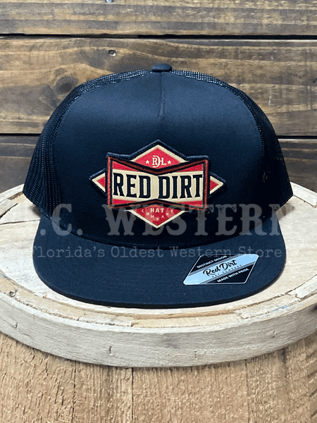 Black trucker hat with Red Dirt cowboy bowtie logo on a wooden barrel front view. If you need any assistance with this item or the purchase of this item please call us at five six one seven four eight eight eight zero one Monday through Saturday 10:00a.m EST to 8:00 p.m EST
