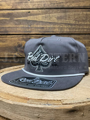 A dark gray snapback cap with a white embroidered spade logo and 'Red Dirt Co.' text on the front, resting on a rustic wooden surface against a wood plank background.If you need any assistance with this item or the purchase of this item please call us at five six one seven four eight eight eight zero one Monday through Saturday 10:00a.m EST to 8:00 p.m EST