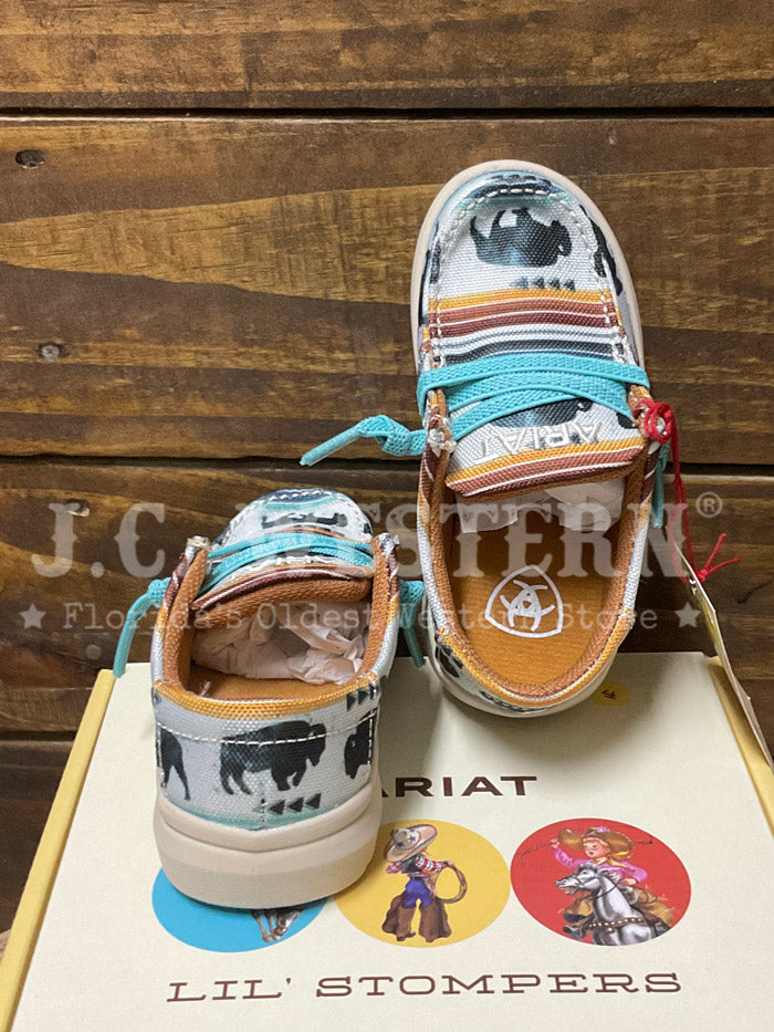 A pair of Ariat slip-on western-style shoes with a multicolor cowboy print and turquoise laces, displayed on top of their yellow branded box against a rustic wooden background. If you need any assistance with this item or the purchase of this item please call us at five six one seven four eight eight eight zero one Monday through Saturday 10:00a.m EST to 8:00 p.m EST