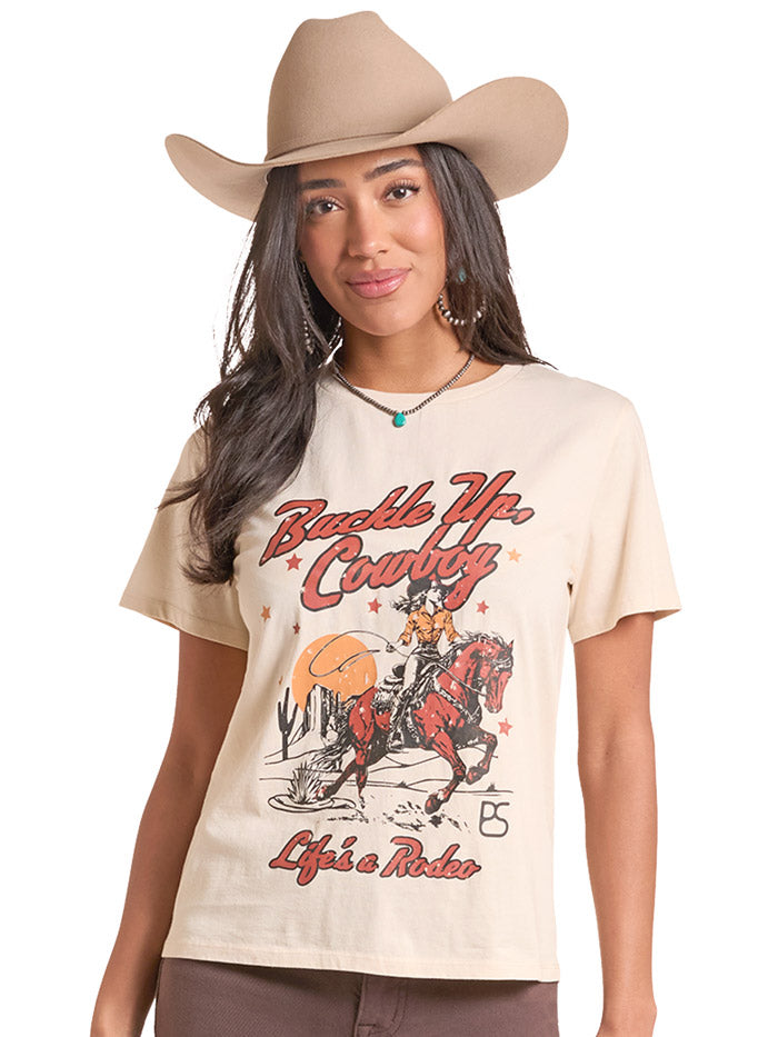 A woman wearing a beige cowboy hat and a cream-colored graphic t-shirt with 'Buckle Up Country' and 'Life's a Rider' printed on the front, set against a plain white background. If you need any assistance with this item or the purchase of this item please call us at five six one seven four eight eight eight zero one Monday through Saturday 10:00a.m EST to 8:00 p.m EST