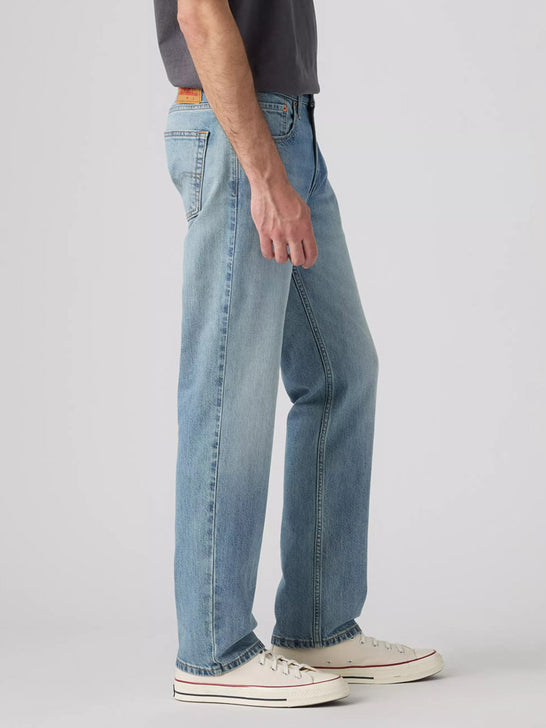 A side profile view of a person wearing light-wash blue denim jeans, a dark gray t-shirt, and white canvas sneakers against a light gray studio background. If you need any assistance with this item or the purchase of this item please call us at five six one seven four eight eight eight zero one Monday through Saturday 10:00a.m EST to 8:00 p.m EST