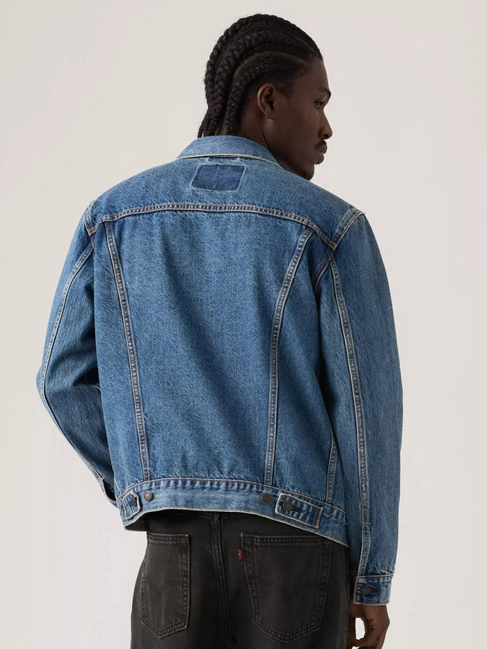 A man wearing a classic medium-wash blue denim jacket over a dark gray t-shirt and black pants, standing against a plain light gray studio background. If you need any assistance with this item or the purchase of this item please call us at five six one seven four eight eight eight zero one Monday through Saturday 10:00a.m EST to 8:00 p.m EST