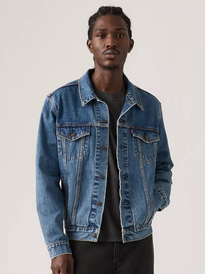 A man wearing a classic medium-wash blue denim jacket over a dark gray t-shirt and black pants, standing against a plain light gray studio background. If you need any assistance with this item or the purchase of this item please call us at five six one seven four eight eight eight zero one Monday through Saturday 10:00a.m EST to 8:00 p.m EST