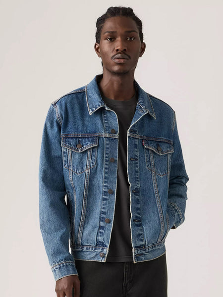 A man wearing a classic medium-wash blue denim jacket over a dark gray t-shirt and black pants, standing against a plain light gray studio background. If you need any assistance with this item or the purchase of this item please call us at five six one seven four eight eight eight zero one Monday through Saturday 10:00a.m EST to 8:00 p.m EST