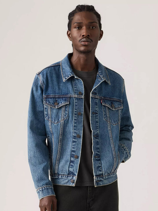 A man wearing a classic medium-wash blue denim jacket over a dark gray t-shirt and black pants, standing against a plain light gray studio background. If you need any assistance with this item or the purchase of this item please call us at five six one seven four eight eight eight zero one Monday through Saturday 10:00a.m EST to 8:00 p.m EST