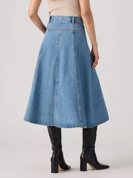 A model shown from the back wearing a high-waisted, light blue denim A-line midi skirt paired with black heeled boots against a light gray studio background. If you need any assistance with this item or the purchase of this item please call us at five six one seven four eight eight eight zero one Monday through Saturday 10:00a.m EST to 8:00 p.m EST