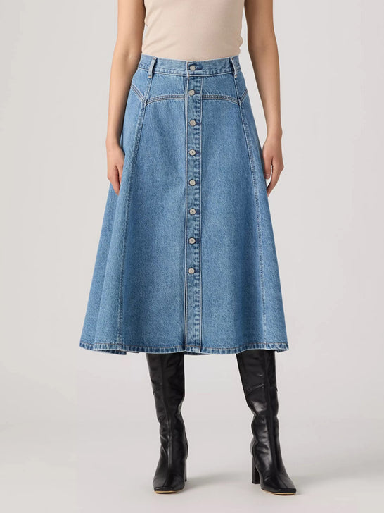 A model wearing a high-waisted, light blue denim A-line midi skirt with a button-down front, paired with a beige top and black leather knee-high boots, against a light gray studio background. If you need any assistance with this item or the purchase of this item please call us at five six one seven four eight eight eight zero one Monday through Saturday 10:00a.m EST to 8:00 p.m EST