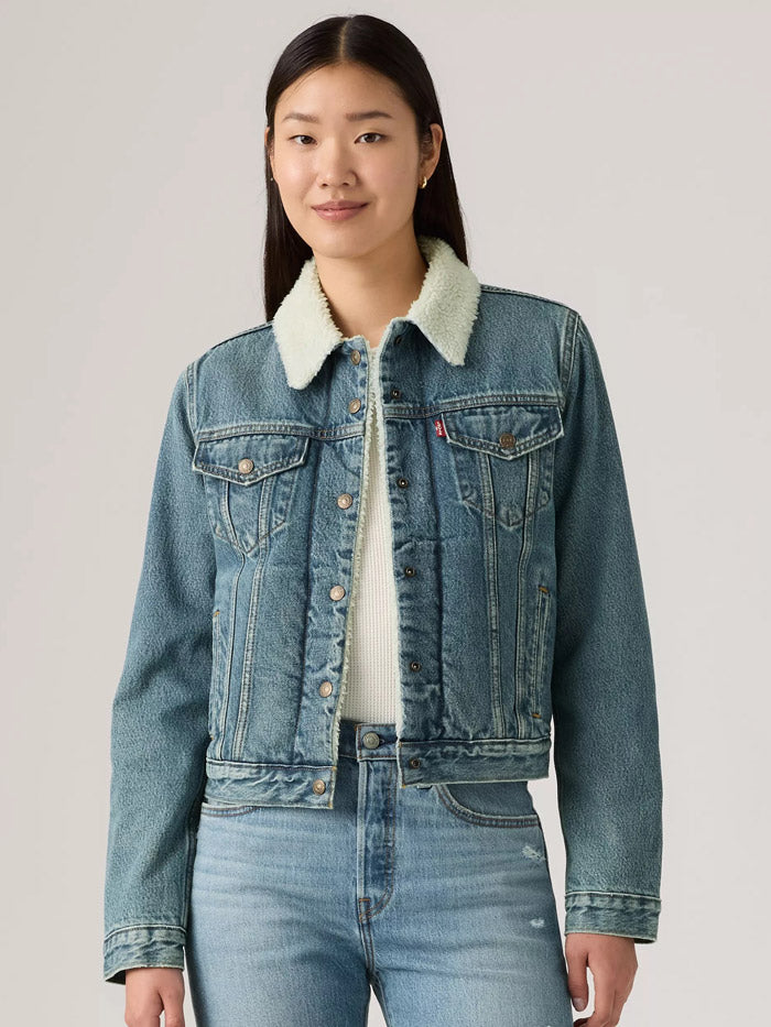A woman modeling a light-wash blue denim jacket with a white sherpa collar, worn over a white top and blue jeans, against a solid light gray background. If you need any assistance with this item or the purchase of this item please call us at five six one seven four eight eight eight zero one Monday through Saturday 10:00a.m EST to 8:00 p.m EST