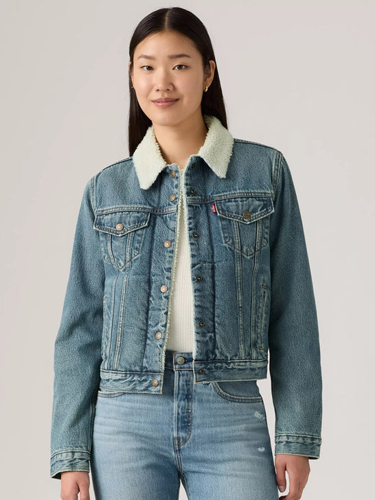 A woman modeling a light-wash blue denim jacket with a white sherpa collar, worn over a white top and blue jeans, against a solid light gray background. If you need any assistance with this item or the purchase of this item please call us at five six one seven four eight eight eight zero one Monday through Saturday 10:00a.m EST to 8:00 p.m EST