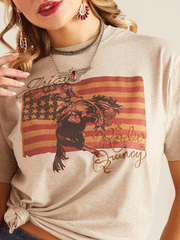 Ariat 10048669 Womens Flag Rodeo Quincy T-Shirt Gold Tan close up view of front graphic. If you need any assistance with this item or the purchase of this item please call us at five six one seven four eight eight eight zero one Monday through Saturday 10:00a.m EST to 8:00 p.m EST