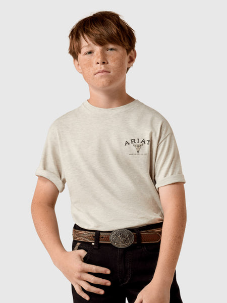 A young boy with red hair and freckles wearing a light beige heathered t-shirt with a black 'Ariat' logo on the left chest, paired with black jeans and a brown leather belt with a silver buckle, against a solid light gray background. If you need any assistance with this item or the purchase of this item please call us at five six one seven four eight eight eight zero one Monday through Saturday 10:00a.m EST to 8:00 p.m EST
